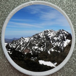 Op de afbeelding: Ronde patch met een bergketen met besneeuwde toppen onder een blauwe lucht. De afbeelding is omlijst door een witte rand. De bergketen is donker met witte sneeuwvlekken.