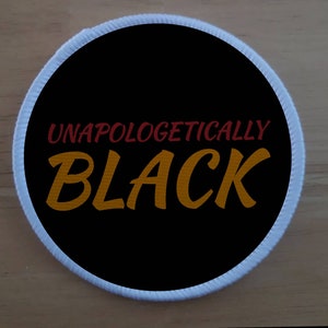 Puede incluir: Un parche redondo negro con costuras blancas alrededor del borde. El texto "Unapologetically Black" está bordado en rojo y dorado.