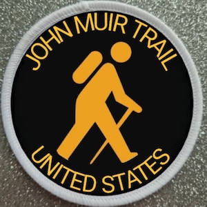 May include: A white and black circular patch with the text "JOHN MUIR TRAIL UNITED STATES" in yellow lettering. A yellow silhouette of a hiker with a backpack and walking stick is in the center of the patch.