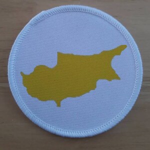 May include: A white circular patch with a yellow outline of the island of Cyprus.