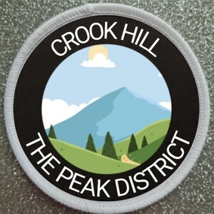 Op de afbeelding: Een witte en zwarte ronde patch met de tekst "Crook Hill The Peak District" en een cartoon afbeelding van een bergketen met een zon en wolken in de lucht.