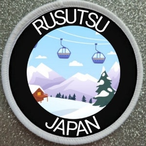 Pode incluir: Um remendo circular branco e preto com o texto "RUSUTSU JAPAN" em letras brancas. O centro do remendo apresenta uma ilustração colorida de uma paisagem montanhosa nevada com uma cabana, um pinheiro e um teleférico com gôndolas.