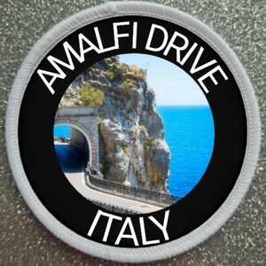 Op de afbeelding: Een ronde zwart-witte patch met de tekst "Amalfi Drive Italy" in wit. De patch toont een foto van een kronkelende weg langs een klif met uitzicht op de oceaan.