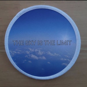 Könnte beinhalten: Ein runder weißer Patch mit einem Bild von blauem Himmel und weißen Wolken. Der Text "THE SKY IS THE LIMIT" ist in Weiß auf dem Patch gedruckt.