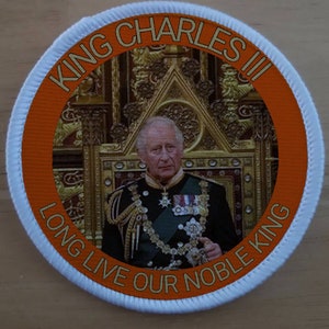 Könnte beinhalten: Ein runder orangefarbener und weißer Aufnäher mit dem Text "King Charles III Long Live Our Noble King" um ein Foto von König Charles III in seinen Krönungsroben.