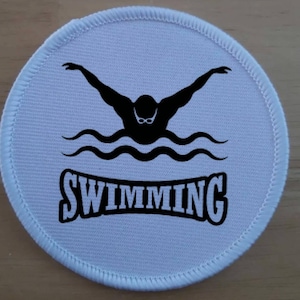 Puede incluir: Parche redondo de tela blanca con costuras negras alrededor del borde. El parche presenta una silueta negra de un nadador con gafas, nadando sobre líneas onduladas negras. La palabra "SWIMMING" está escrita en letras negras debajo del nadador.