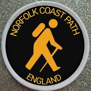 May include: A round, white-bordered patch with a black background. The patch features a yellow silhouette of a hiker with a backpack and walking stick. The text "NORFOLK COAST PATH ENGLAND" is written in yellow around the hiker.