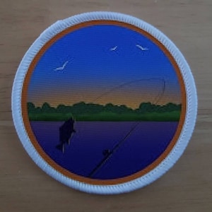 Puede incluir: Un parche redondo blanco con un borde naranja. El parche presenta una imagen escénica de un lago al atardecer con una caña de pescar y un pez saltando fuera del agua. También hay pájaros volando en el cielo.