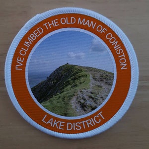 Puede incluir: Parche redondo con borde naranja y costuras blancas. El parche presenta una imagen de paisaje de una montaña verde con un sendero. El texto en el parche dice "I'VE CLIMBED THE OLD MAN OF CONISTON" y "LAKE DISTRICT".
