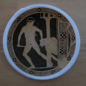May include: Circular patch featuring a black and beige illustration of a classical scene. A figure with a sword confronts a beast, set against columns and a decorative border. The patch has a white edge.