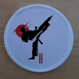 May include: White round patch featuring a black silhouette of a person performing a karate kick against a red sun. The text "空手" is written in red beneath the figure.