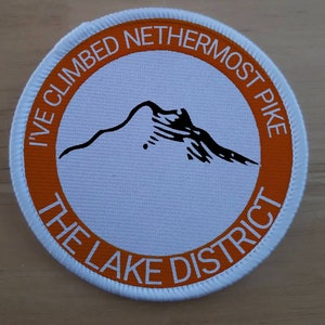 May include: A white and orange round patch with the text "I've Climbed Nethermost Pike The Lake District" and a black silhouette of a mountain.