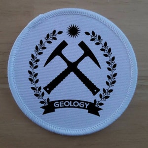 Puede incluir: Parche redondo blanco con bordado negro. El diseño presenta dos picos cruzados, una corona de hojas, un sol con rayos y la palabra "GEOLOGÍA" en una pancarta.