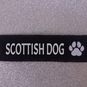 May include: Black rectangular patch with the words "SCOTTISH DOG" in white capital letters. A white paw print is on the right side. The patch is on a sparkly silver background.