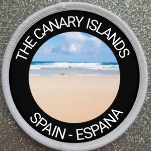 Può includere: Toppa rotonda bianca con bordo nero e il testo "THE CANARY ISLANDS SPAIN - ESPANA" attorno a una foto di una spiaggia con cielo blu e nuvole bianche.