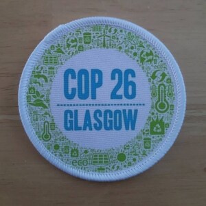May include: A round white patch featuring the text "COP 26 GLASGOW" in blue. The patch is encircled by a border of green icons representing environmental themes. The patch is on a wooden surface. The patch is a symbol of the COP26 climate summit.