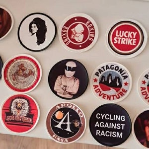 May include: A collection of 13 round embroidered patches with various designs. The patches feature images of a woman, a car, a globe, a rocket, a mountain range, a butterfly, a cigarette, a star, and text. The patches are all different colours and have a variety of designs.