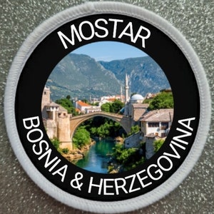 May include: A round patch with a black background and white text. The text reads "Mostar Bosnia & Herzegovina". The center of the patch features a photo of a bridge over a river with a city in the background.