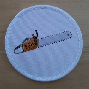 Peut inclure: Un patch rond en tissu blanc avec un motif de tronçonneuse noir et orange. La tronçonneuse a une poignée noire et une lame argentée avec une bordure noire dentelée.