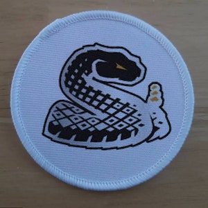Puede incluir: Parche redondo blanco con un diseño de serpiente en blanco y negro. La serpiente está enroscada con la cabeza mirando hacia la izquierda. La serpiente tiene un ojo dorado.