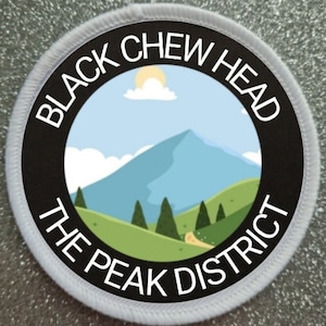Può includere: Un cerotto circolare bianco e nero con il testo "Black Chew Head The Peak District" in un cerchio nero con una scena di montagna di cartone animato all'interno. La montagna è blu e verde con una nuvola bianca e un sole.