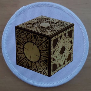 May include: A white round patch with a gold and brown cube with intricate designs on all sides. The cube is a replica of the iconic Tesseract from the movie "Avengers: Endgame".