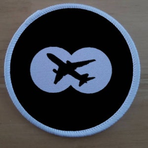 Könnte beinhalten: Ein runder schwarzer und weißer Patch mit einer weißen Flugzeugsilhouette in zwei weißen Kreisen. Der Patch hat einen weißen Rand.