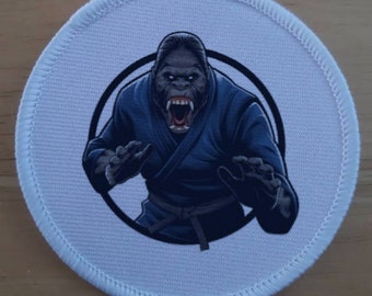 May include: A round patch featuring a gorilla in a blue gi, with an open mouth and aggressive stance. The gorilla is inside a black circle, set against a light blue background with a white stitched border. The patch is designed for martial arts enthusiasts.