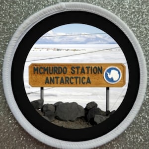 May include: A white and black round patch with a black circle in the center. The circle shows a sign that reads "MCMURDO STATION ANTARCTICA" with a blue circle with a white outline and a white penguin inside. The sign is in front of a snowy landscape with mountains in the distance.