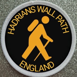 May include: A black and white round patch with a white border. The patch features a yellow silhouette of a hiker with a backpack and walking stick. The text "Hadrian's Wall Path England" is written in yellow around the hiker.