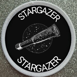May include: A black and white embroidered patch with the text "STARGAZER" in a circle around a telescope pointing towards a circle of stars. The patch has a white border.