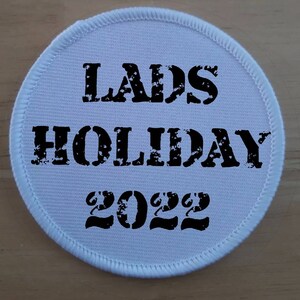 Könnte beinhalten: Runder weißer Aufnäher mit schwarzem Text, der "LADS HOLIDAY 2022" lautet.