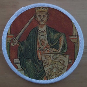 May include: A round embroidered patch featuring a medieval-style illustration of a king seated on a throne, holding a sword and a shield with three lions. The king is wearing a crown and a green robe. The background is red with gold accents.