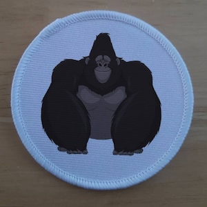 Può includere: Una toppa rotonda blu chiaro con un'illustrazione di un gorilla dei cartoni animati. Il gorilla è nero con il petto grigio e un'espressione sorridente. La toppa ha un bordo bianco cucito.