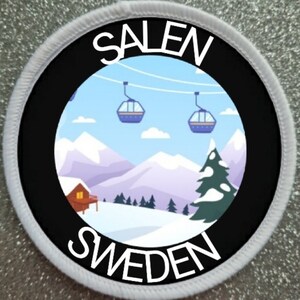May include: A white circular patch with black text and a black circle in the centre. The text reads "SALEN SWEDEN". Inside the black circle is a colourful illustration of a snowy mountain scene with ski lifts and a small cabin.
