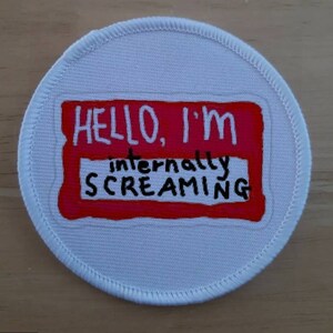 May include: A white round patch with a red name tag that says "HELLO, I'M internally SCREAMING".