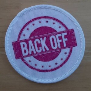 May include: White round fabric patch with pink stitching around the edge. The patch has a pink circle with white stars around the edge and the words "BACK OFF" in white letters inside the circle.