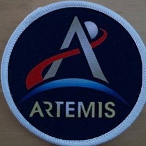 Peut inclure: Écusson rond avec un fond bleu marine et une bordure blanche. Le patch présente le logo Artemis, avec un design en forme de A argenté, un arc rouge et blanc, et le mot ARTEMIS en or.