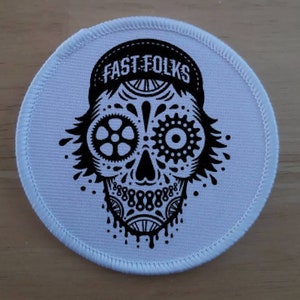 May include: White round patch with a black and white illustration of a skull wearing a baseball cap with the text "FAST FOLKS" on it. The skull's eyes are made of gears.