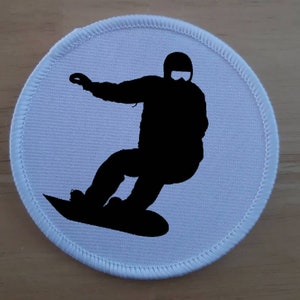 Könnte beinhalten: Runder weißer Patch mit einer schwarzen Silhouette eines Snowboarders auf einem Snowboard.