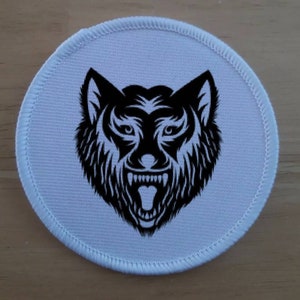 May include: A white round patch with a black and white illustration of a wolf's head with a snarling expression.