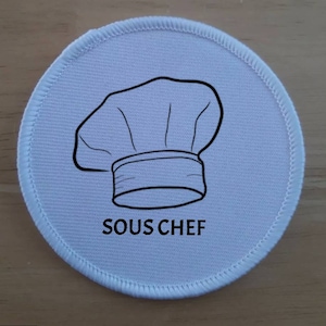 Può includere: Toppa rotonda in tessuto bianco con un contorno nero di un cappello da cuoco e il testo "SOUS CHEF".