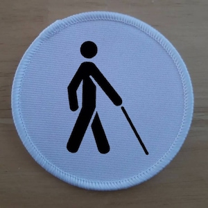 May include: White round fabric patch with a black silhouette of a person walking with a white cane. The patch has a white border with a stitched edge.