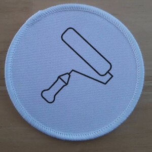 May include: A circular, pale blue patch featuring a black outline of a paint roller. The patch has a white stitched border. The paint roller is illustrated in a simple, graphic style.