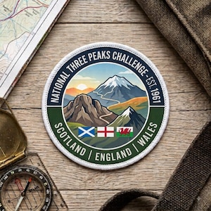 May include: A circular embroidered patch featuring a mountain landscape with the text NATIONAL THREE PEAKS CHALLENGE - EST. 1961. Below the mountains are the flags of Scotland, England, and Wales, with the country names printed along the bottom border.