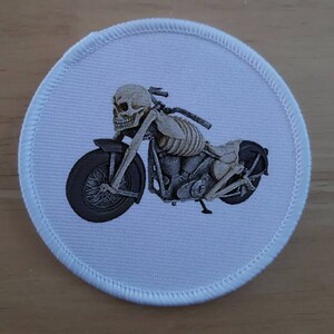 May include: A white round patch with a skeleton riding a motorcycle. The motorcycle is black and chrome with a skull for a head.