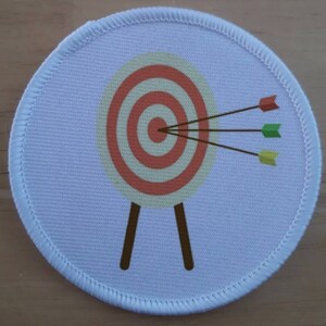 May include: A white fabric patch with a red and white target with three arrows, one red, one green, and one yellow, hitting the target.