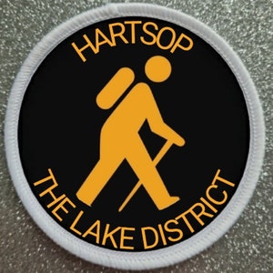 May include: A black and white round patch with a white border. The patch features a yellow stick figure hiker with a backpack and walking stick. The text "HARTSOP THE LAKE DISTRICT" is written in yellow around the hiker.