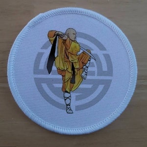 May include: White round patch with a gray circle design in the center. A cartoon image of a person in a yellow and orange robe holding a sword is in the center of the circle.