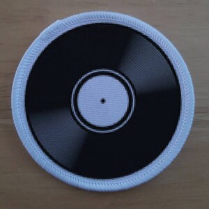 May include: A white vinyl record patch with a black centre and a white label. The patch is surrounded by a white border.